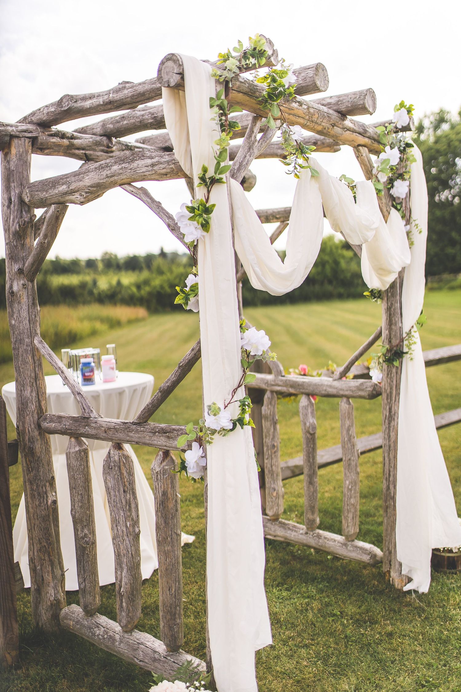 Rustic wedding ceremony arch made of weathered wood and white fabric decorates an outdoor venue.