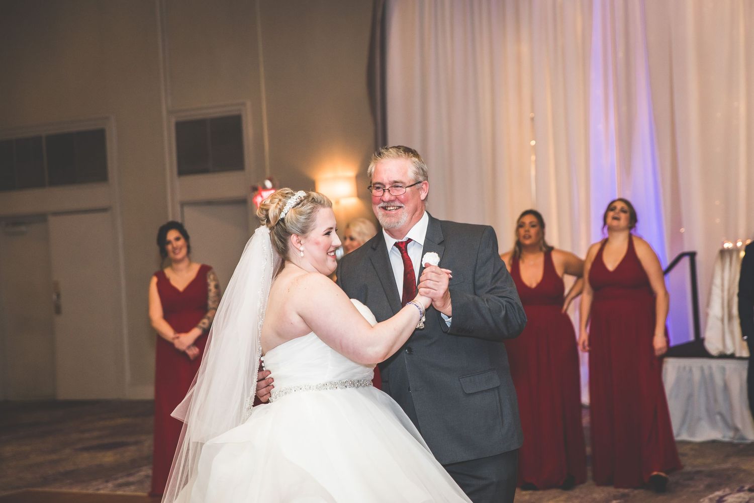 Father-daughter dance at wedding reception with bridesmaids in burgundy dresses watching.