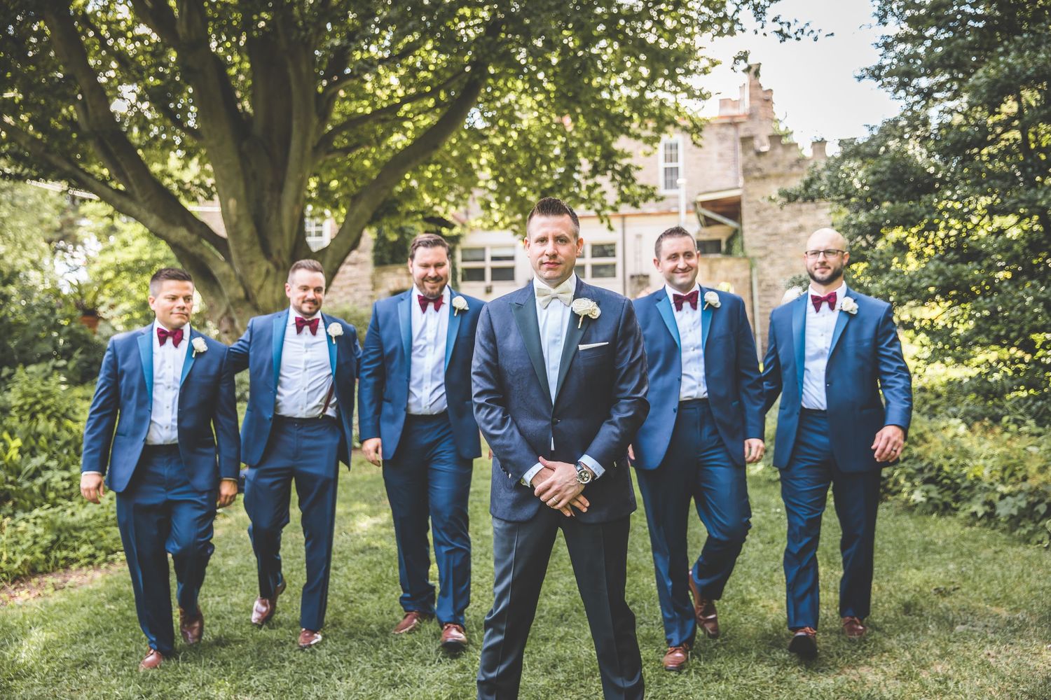 Groomsmen in navy blue suits pose together outdoors for wedding party photos.
