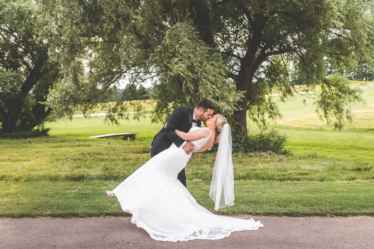 Dramatic dip kiss between newlyweds on a tree-lined path with flowing wedding dress.
