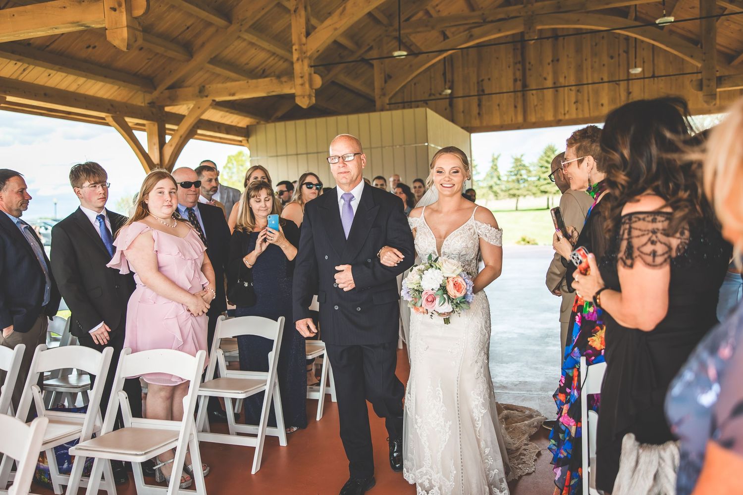 Wedding ceremony processional in a rustic wooden barn venue with guests seated on white chairs.