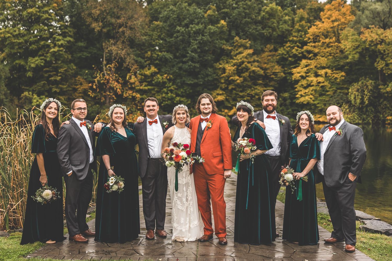 Large wedding party in formal attire stands together on stone path by autumn lake.