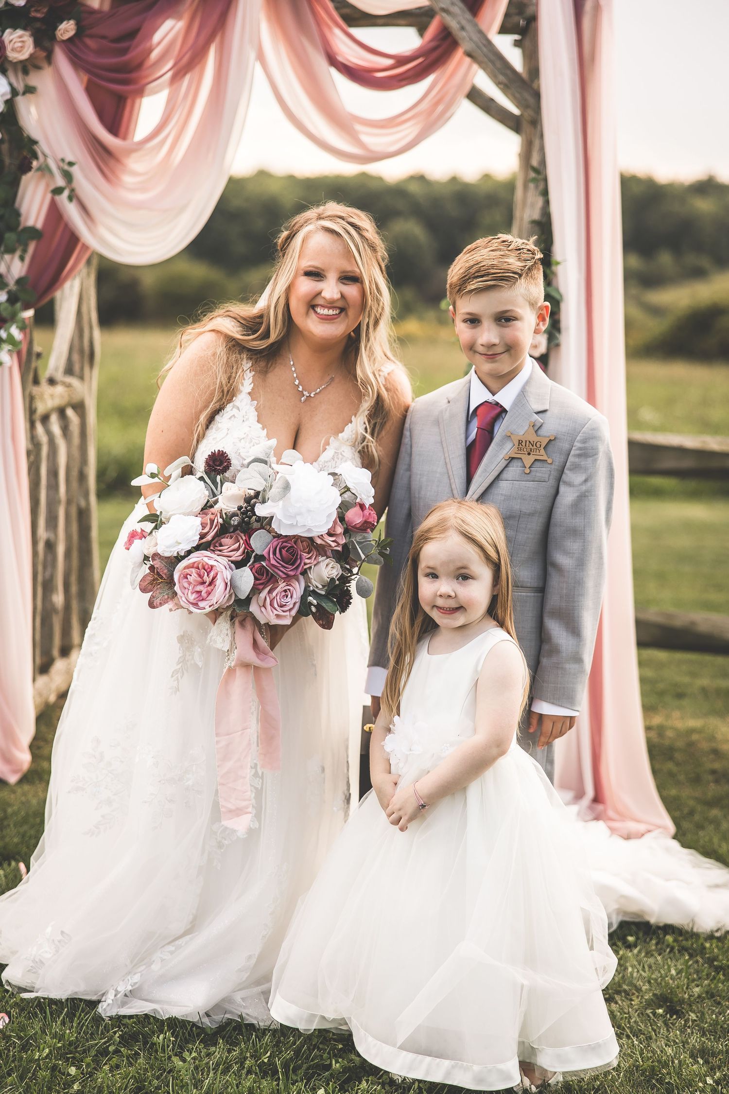 Wedding party poses under pink fabric draped arch with flower girls in white dresses.
