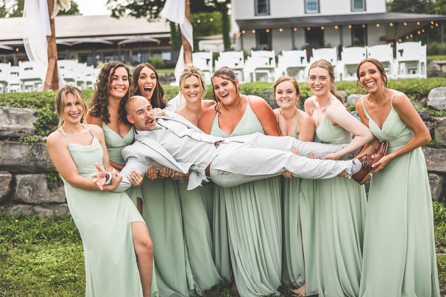 Bridesmaids in mint green dresses playfully carry someone while posing on grassy hill.