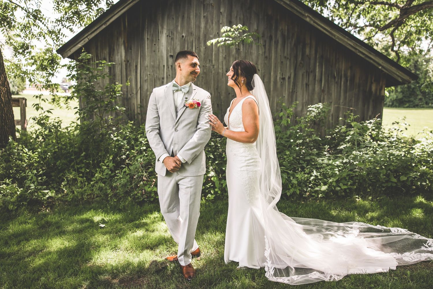 Couple in wedding attire embraces near wooden barn in sunlit garden setting.