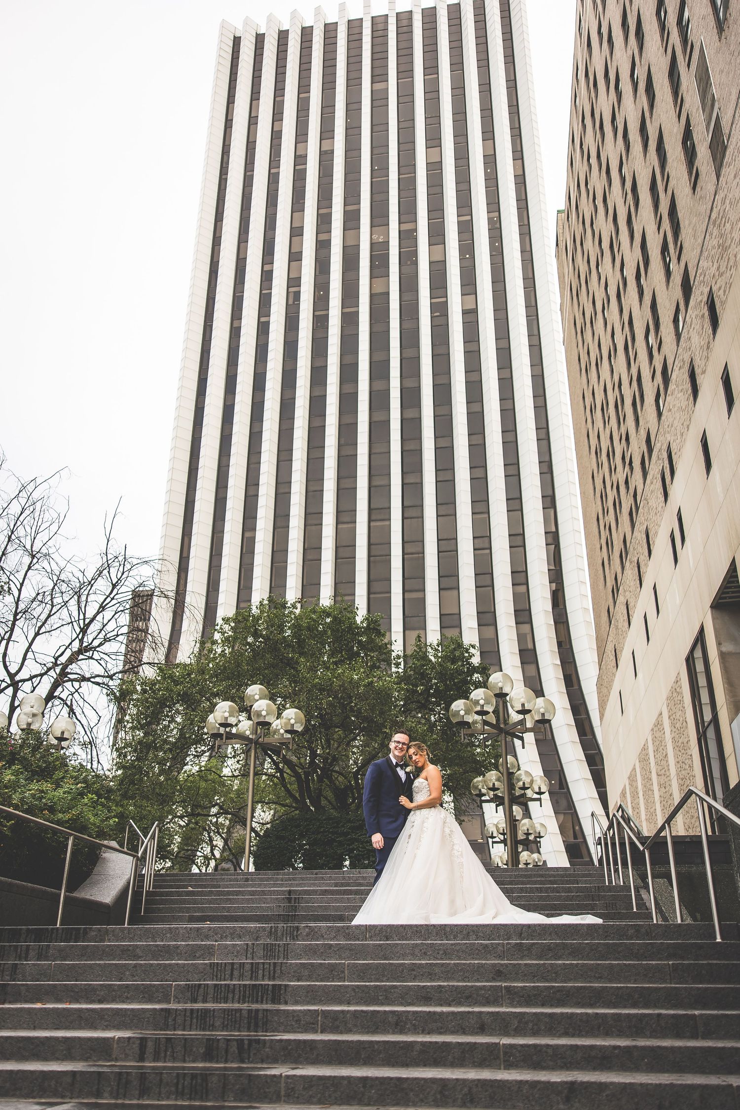 A couple in wedding attire poses on grand stone steps in front of a modern high-rise building.