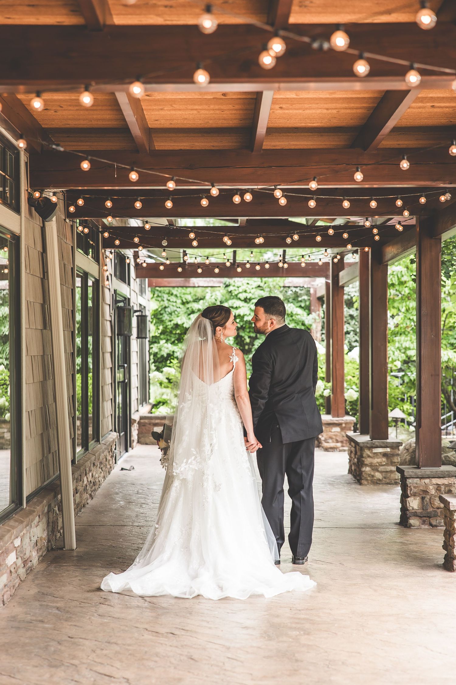 Wedding couple stands under rustic wooden pergola decorated with string lights.