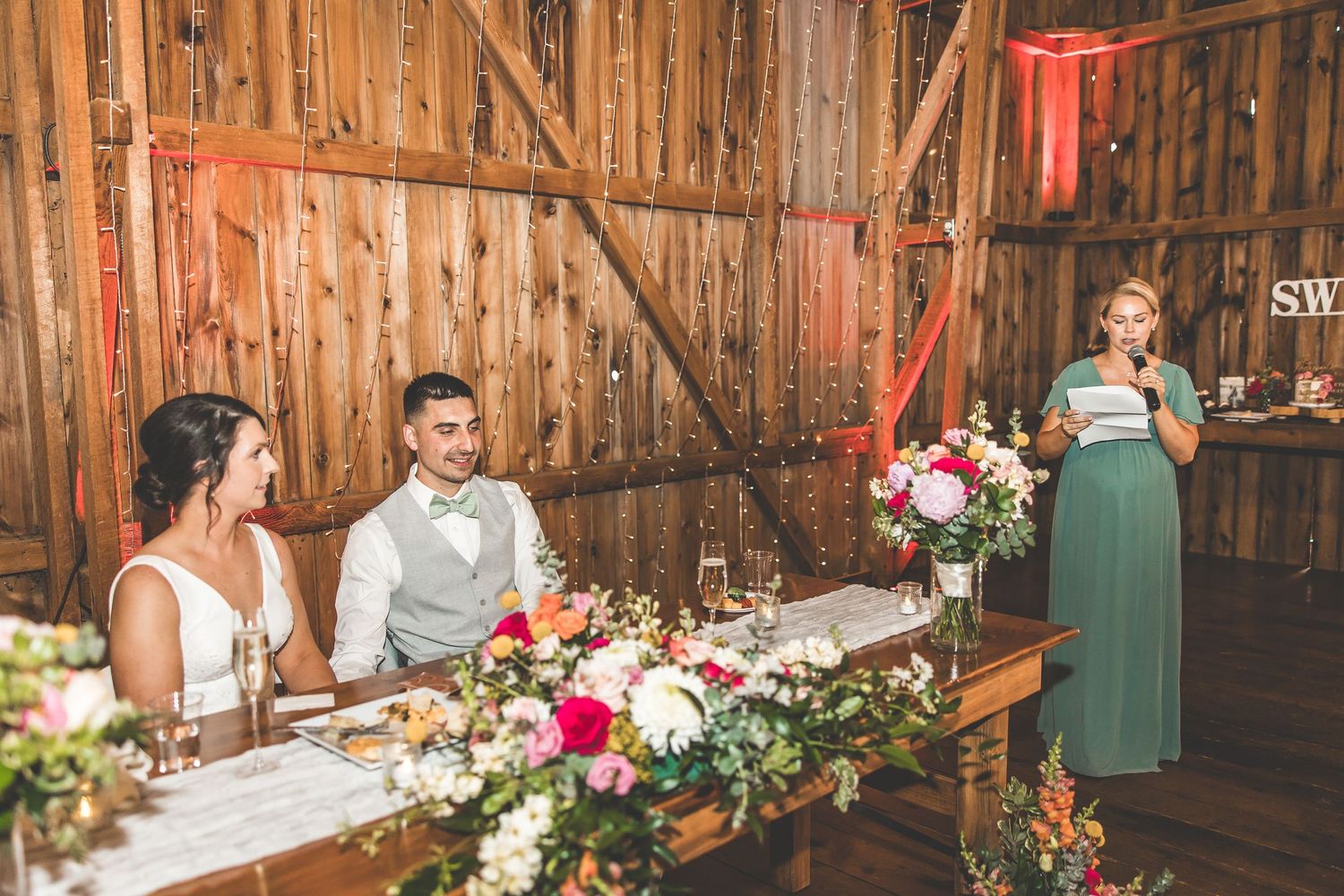 A speech being delivered during a wedding reception in a decorated barn with floral arrangements.