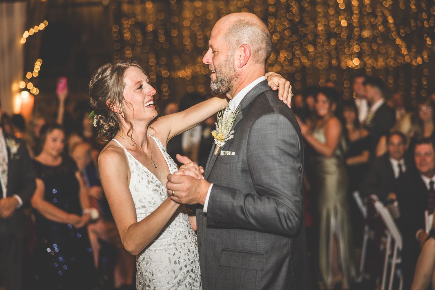 Wedding couple dances beneath string lights in rustic barn reception venue.
