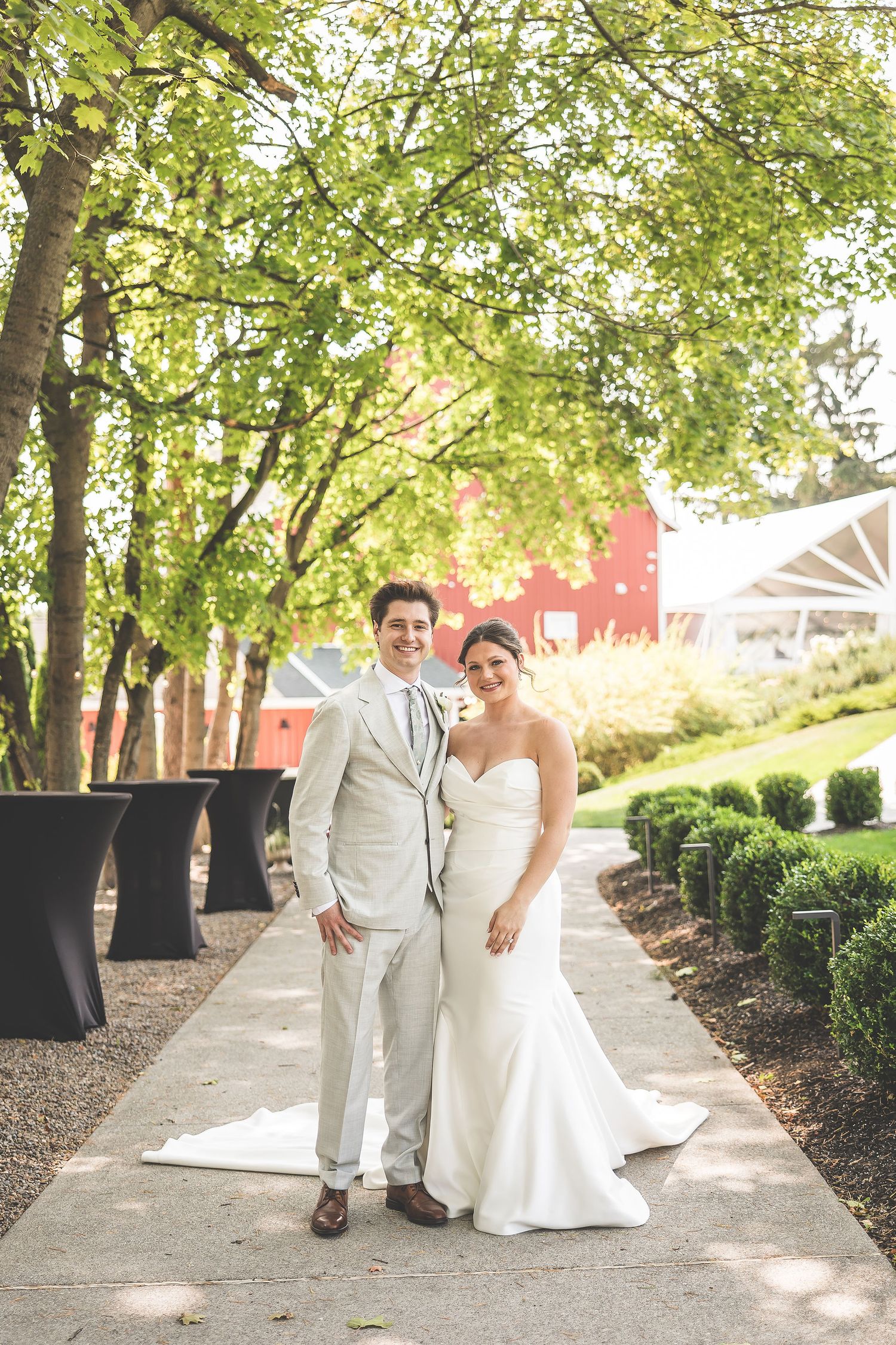 Wedding couple poses along a landscaped pathway with trees and a red barn in the background.