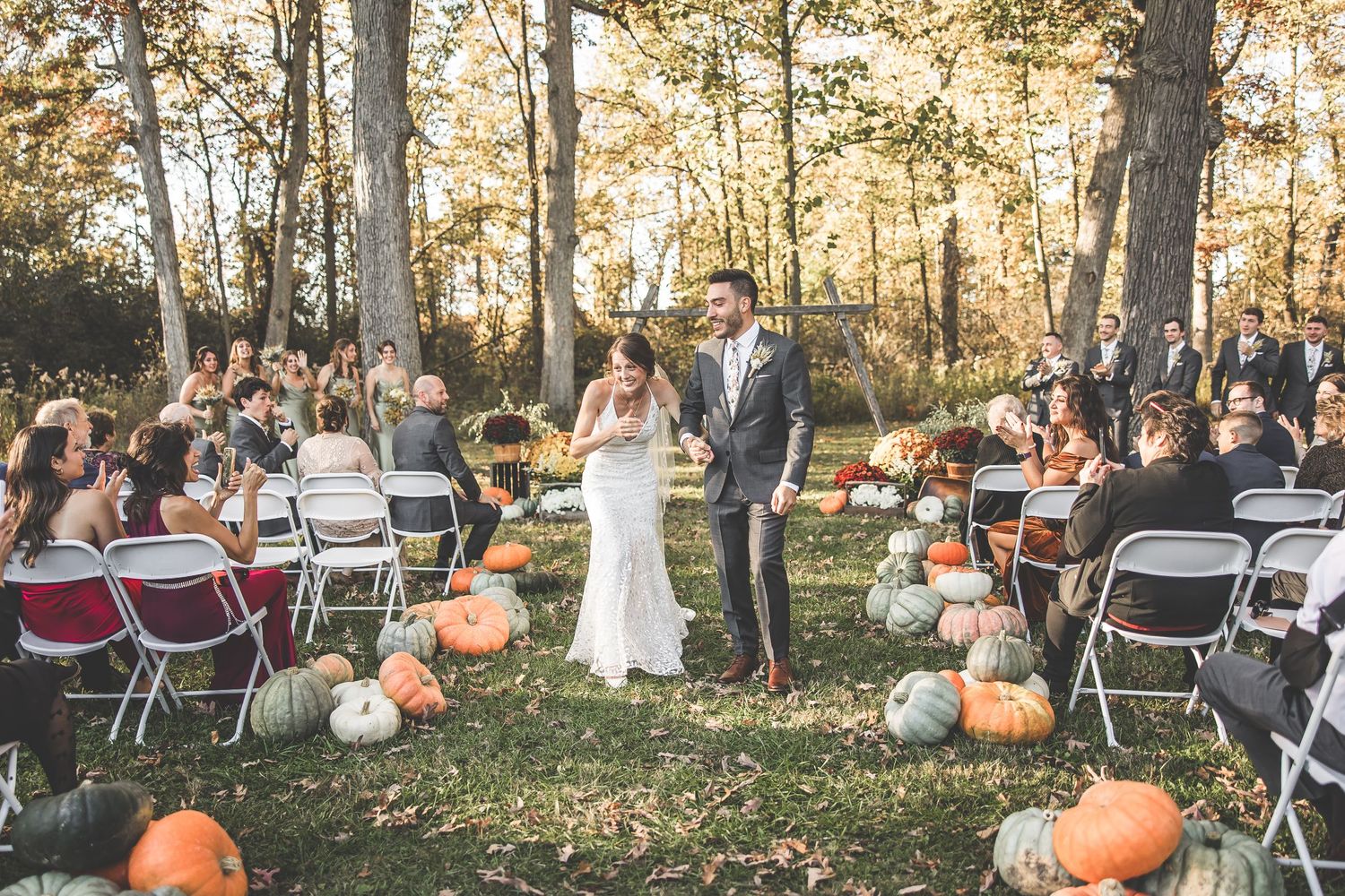 Outdoor wedding ceremony with guests seated on folding chairs among trees and pumpkin decorations.