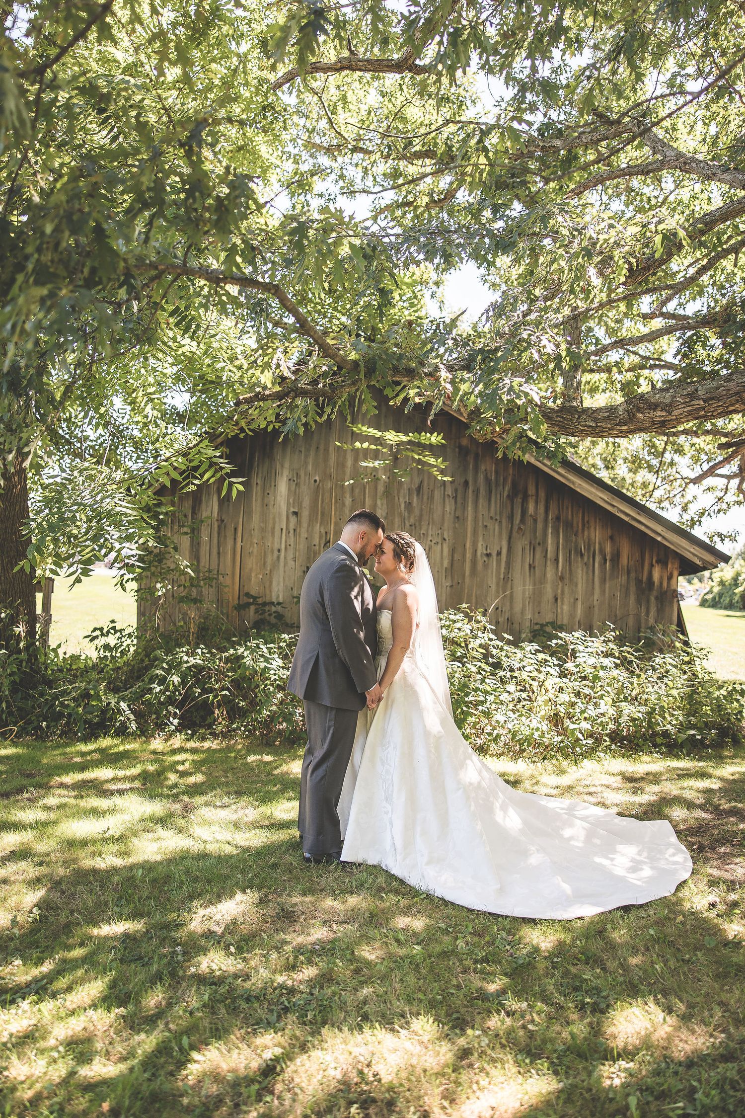 Romantic wedding portraits taken in front of a weathered barn with natural sunlight filtering through trees.