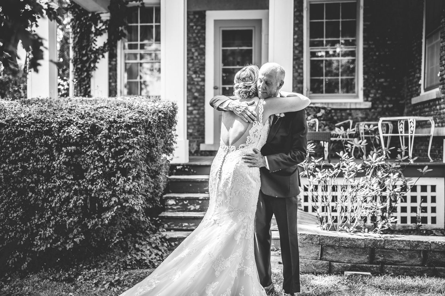 A black and white wedding photo captures a joyful moment between newlyweds outside a brick house.