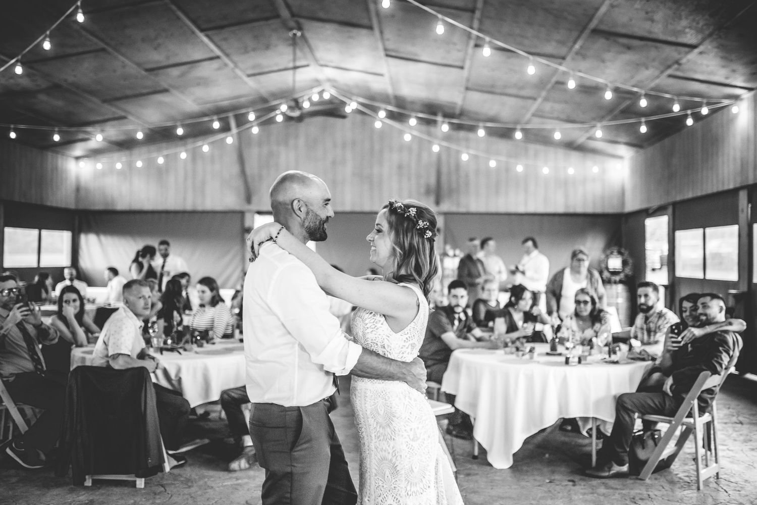 Wedding guests dance together on a reception dance floor under string lights and exposed beams.