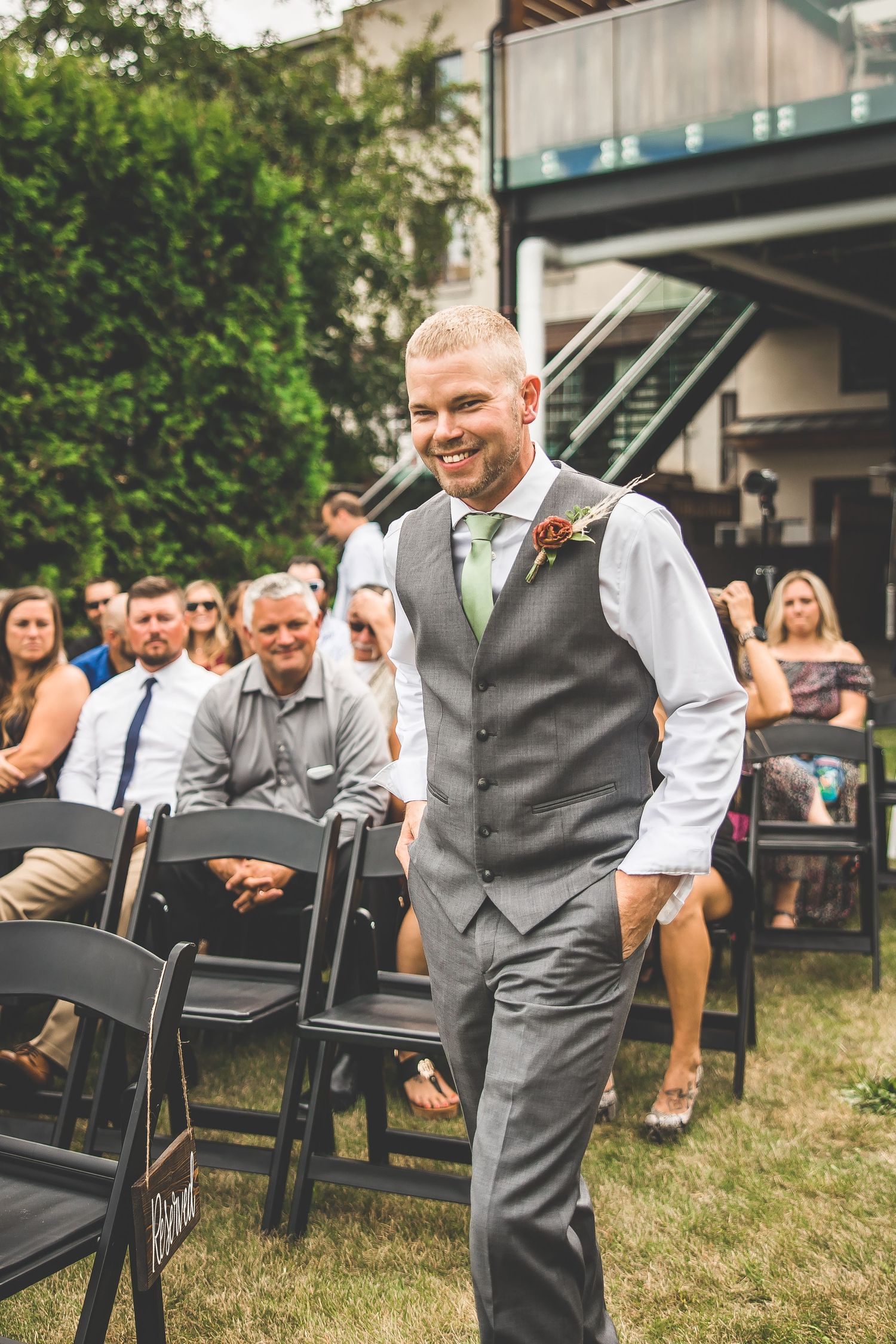 Wedding guests seated in outdoor ceremony area with grey chairs on grass.