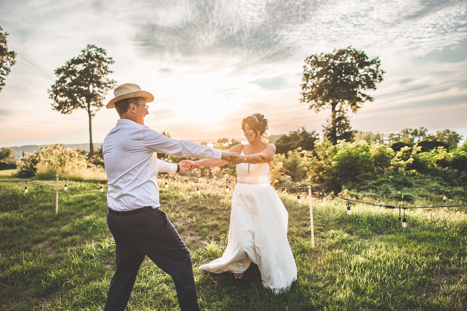 A couple dances playfully in a sunlit field during their wedding celebration.