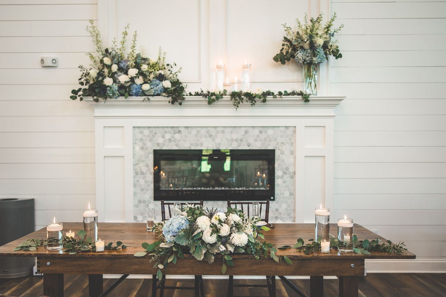 Elegant wedding reception table setup with floral arrangements and candles in front of a white fireplace.
