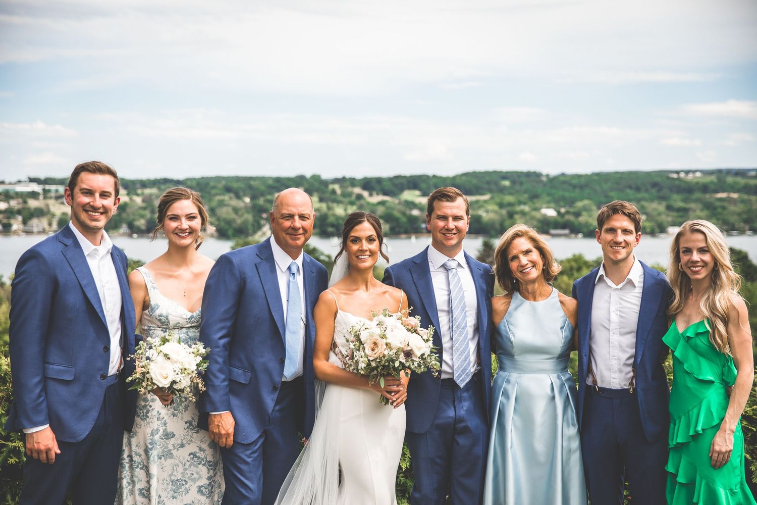 Wedding party in blue and pastel attire poses with scenic lake view background.