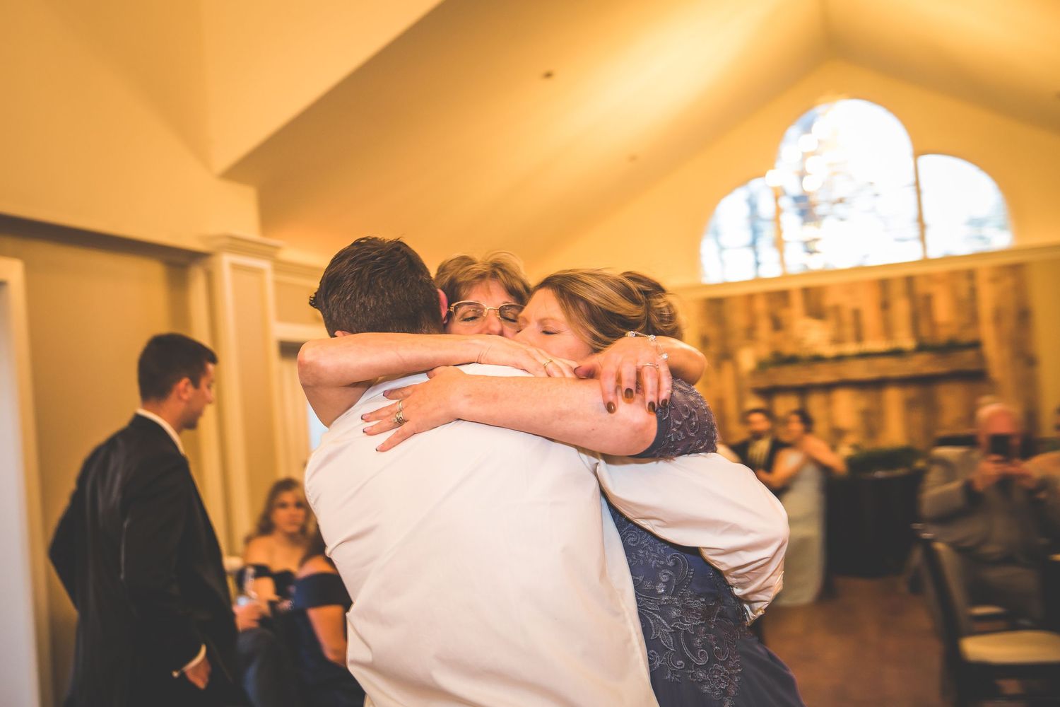 A couple embraces while dancing at a wedding reception with warm lighting and guests in the background.