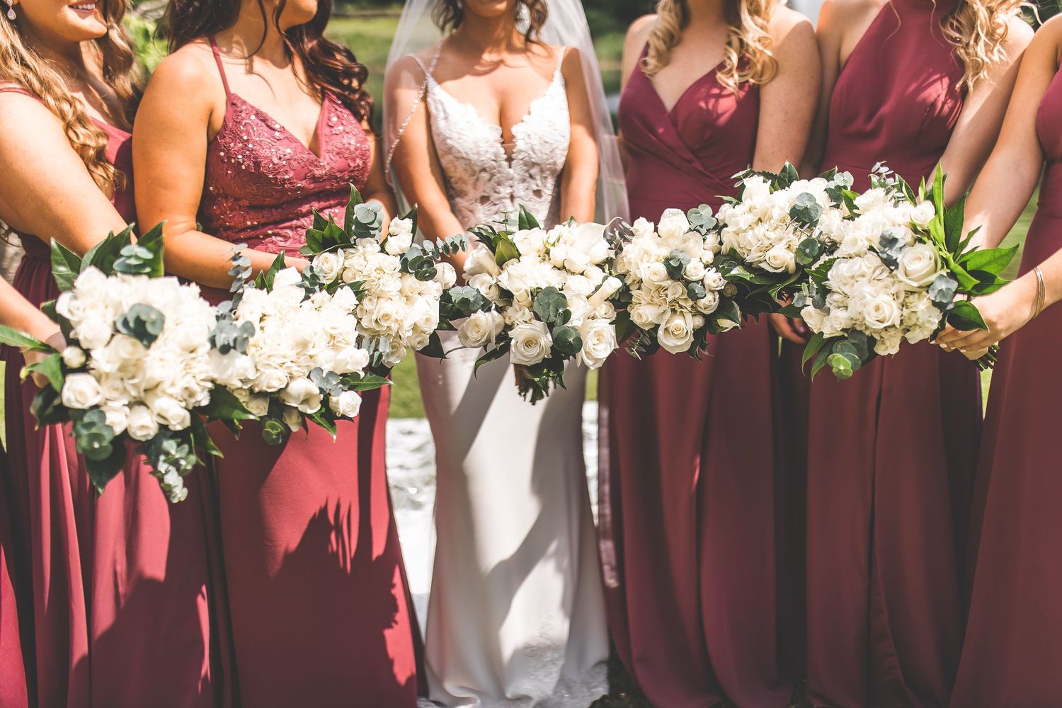 A row of white floral bouquets held by attendants in burgundy dresses at an outdoor wedding.