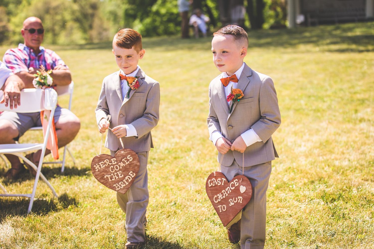 Ring bearers in gray suits carry wooden heart signs during an outdoor ceremony.