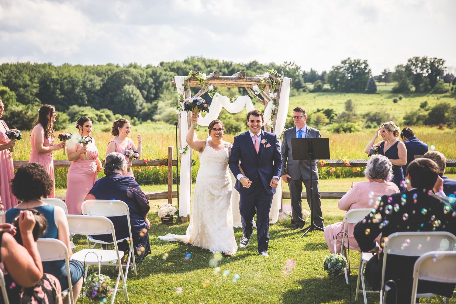 A sunny outdoor wedding ceremony with guests seated in white chairs and attendants in pink dresses.