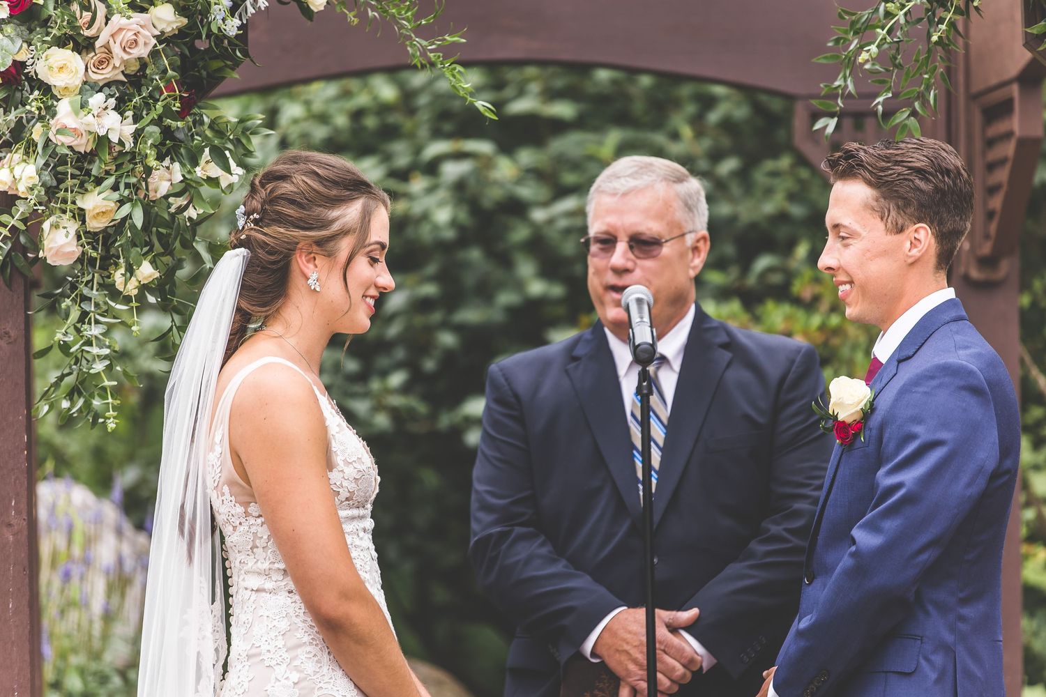 Wedding ceremony participants gathered under wooden pergola with floral arrangements.