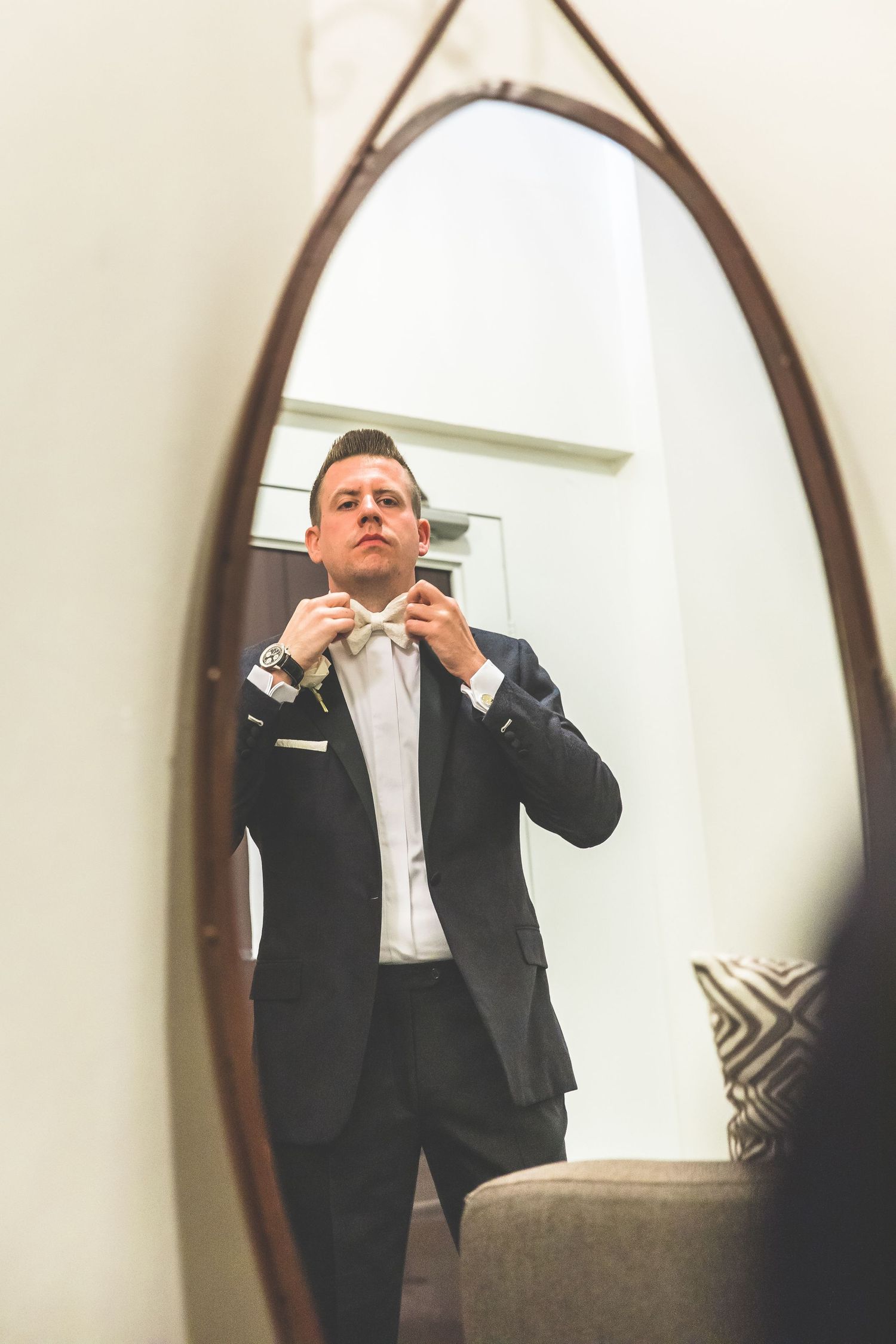 Mirror reflection shows groom adjusting tie before ceremony in arched frame.