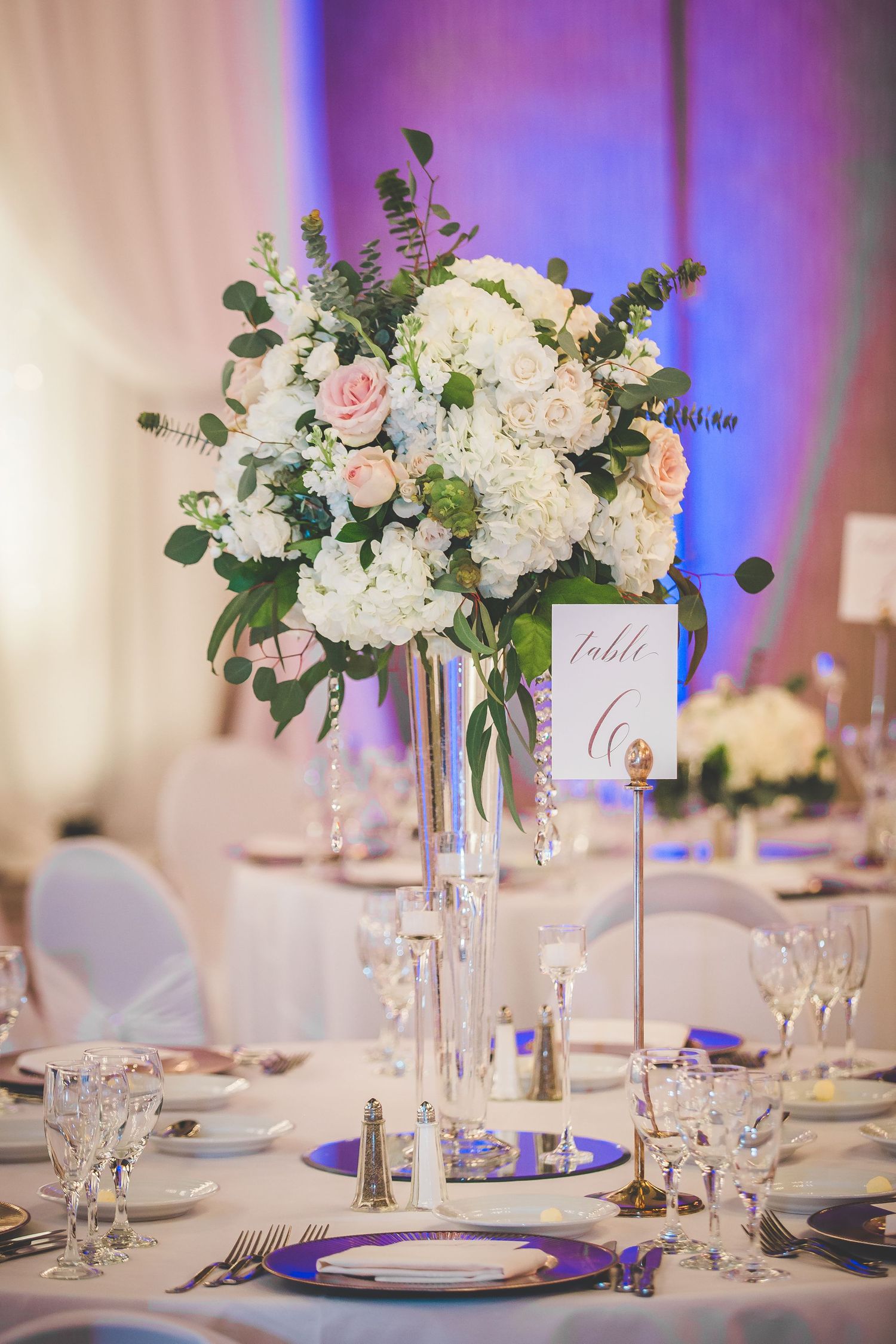 Elegant wedding reception table settings with tall white floral centerpieces and soft purple lighting accents.
