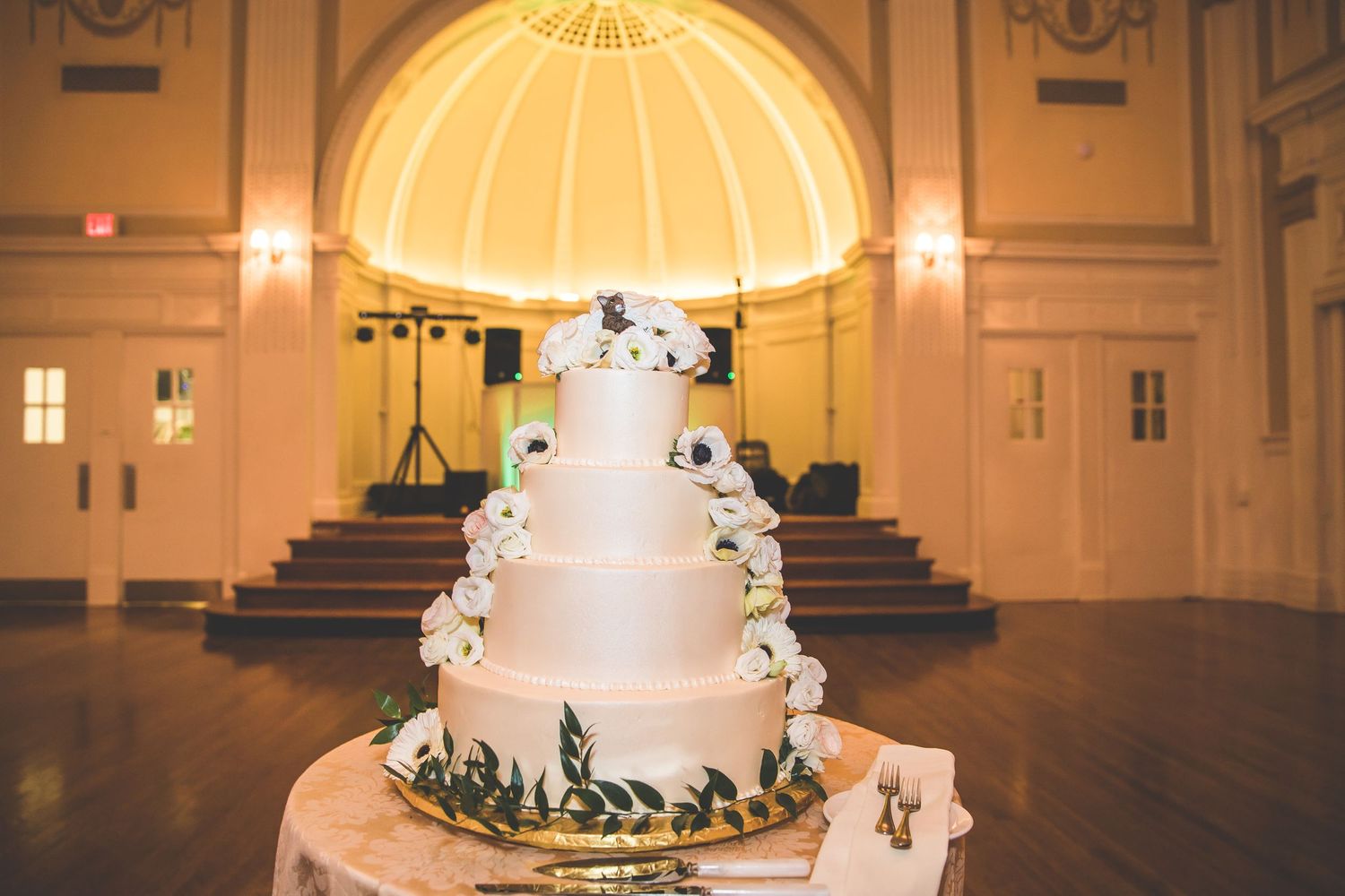 Classic white tiered wedding cake displayed under a golden domed ceiling in an elegant ballroom.