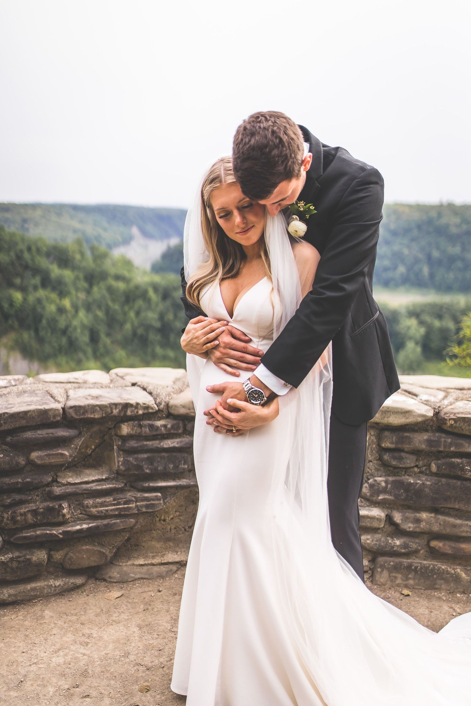 Romantic wedding couple embraces against stone wall overlooking scenic mountain landscape.