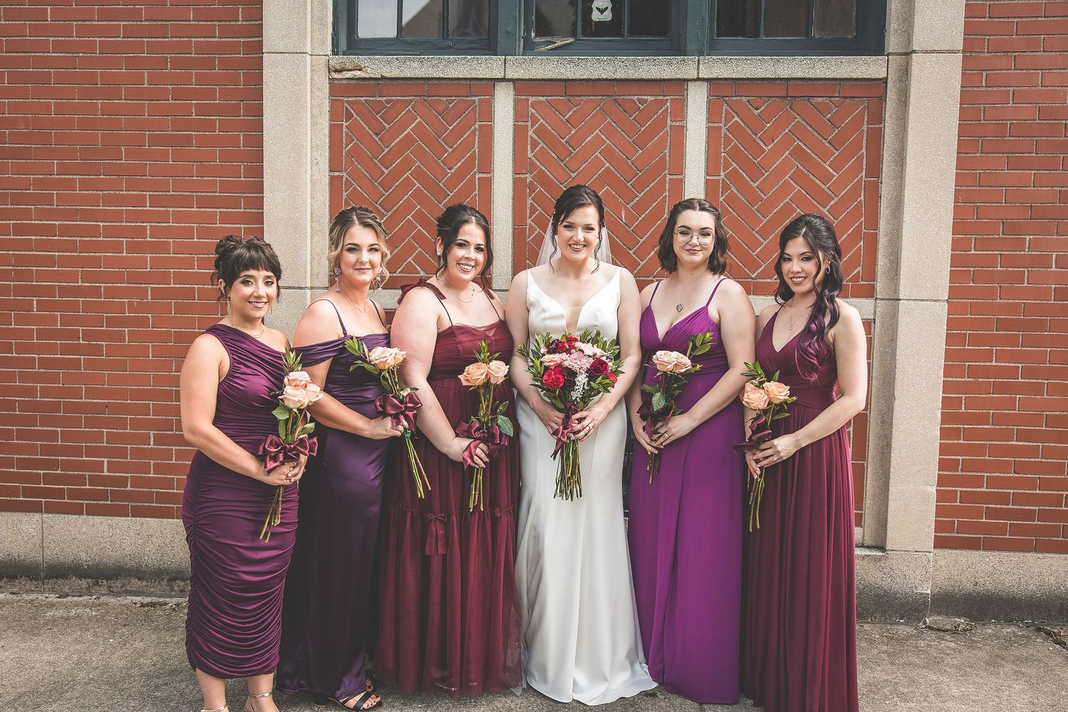 Bridal party in purple dresses posed together against red brick wall.