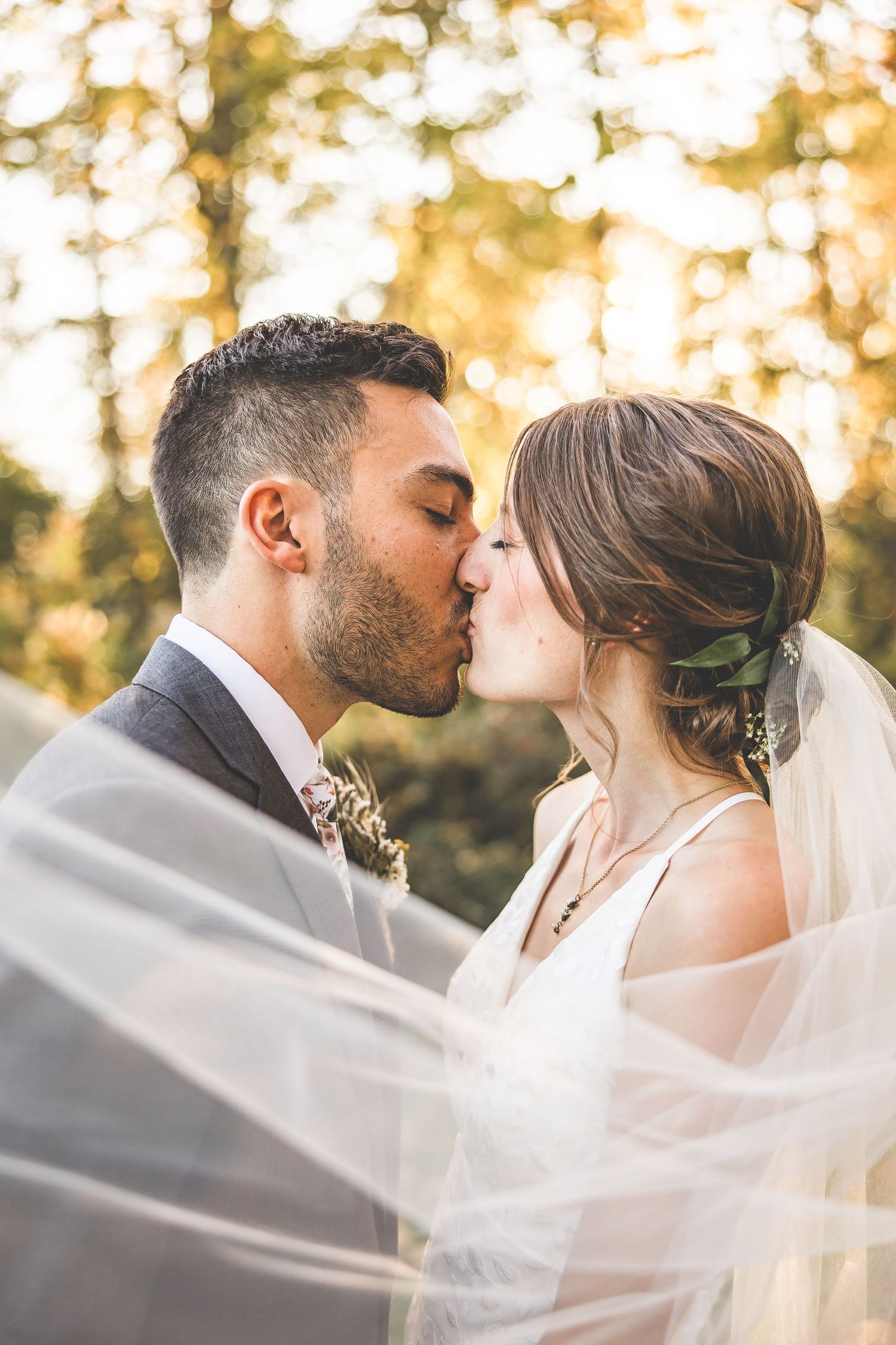 Romantic kiss through flowing veil with golden sunlight in forest background.