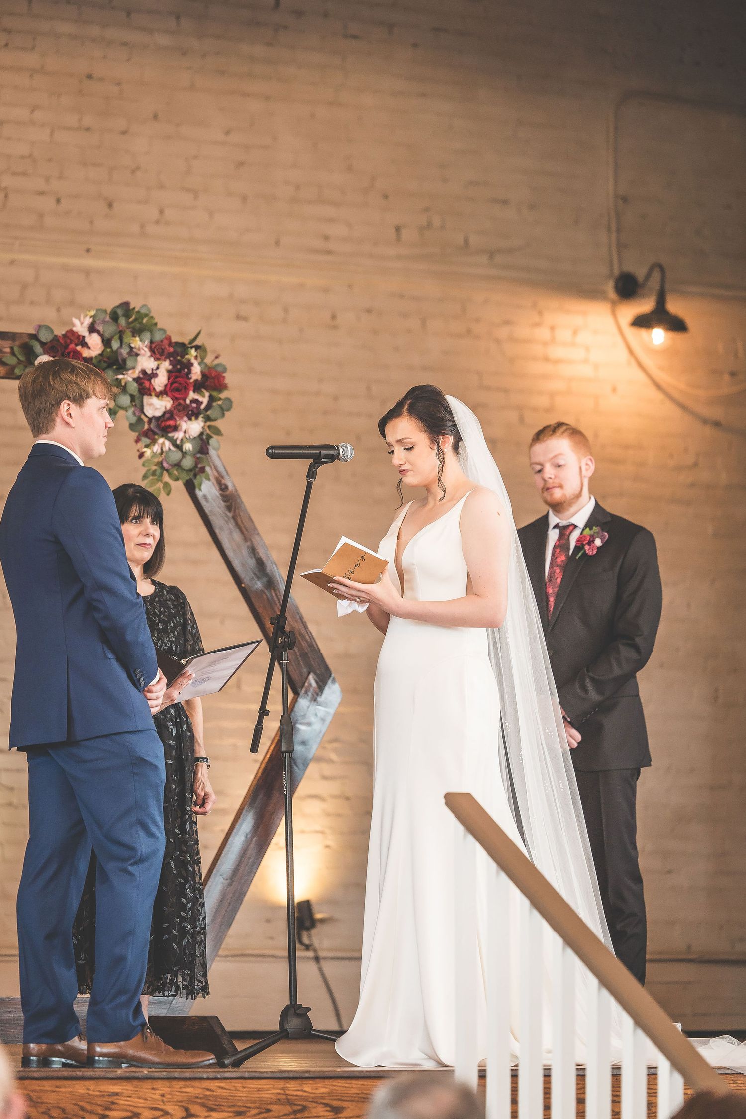 Wedding ceremony taking place in an elegant brick-walled venue with wooden decor and floral arrangements.