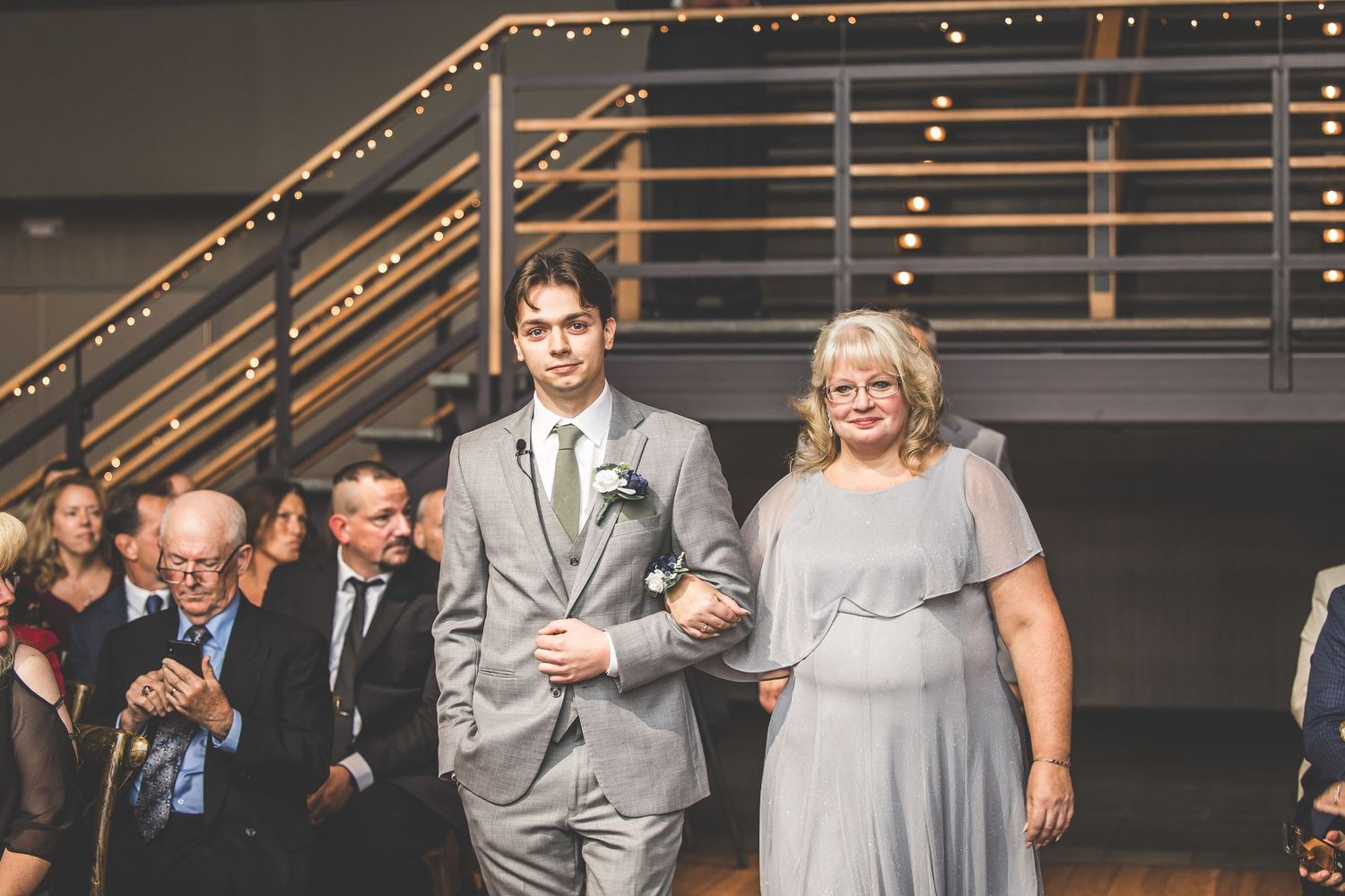 Wedding party members walk down the aisle at a rustic indoor venue with wooden beams and string lights.