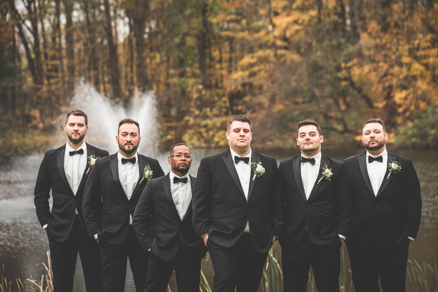Groomsmen in black tuxedos pose together during fall wedding celebration.