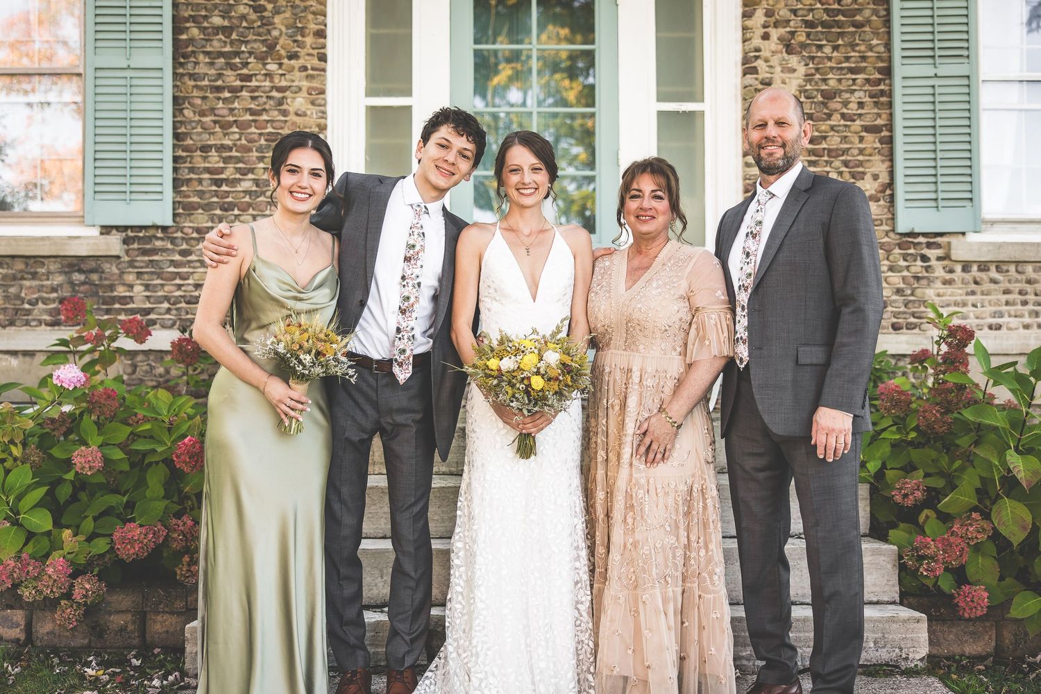 Wedding party poses together on stone porch with mint green shutters and flowering bushes.