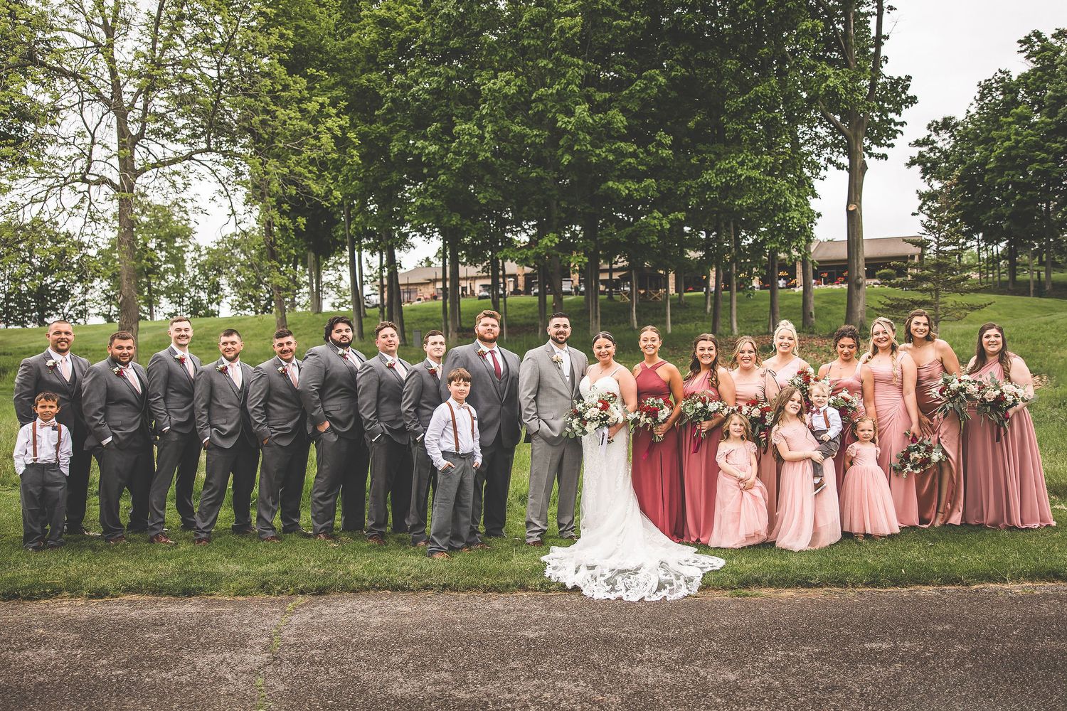 Wedding party in pink dresses and gray suits poses together on a grassy hillside.
