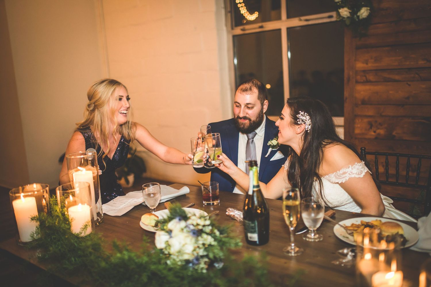 Wedding reception dinner setting with candles and floral arrangements on rustic wooden tables.