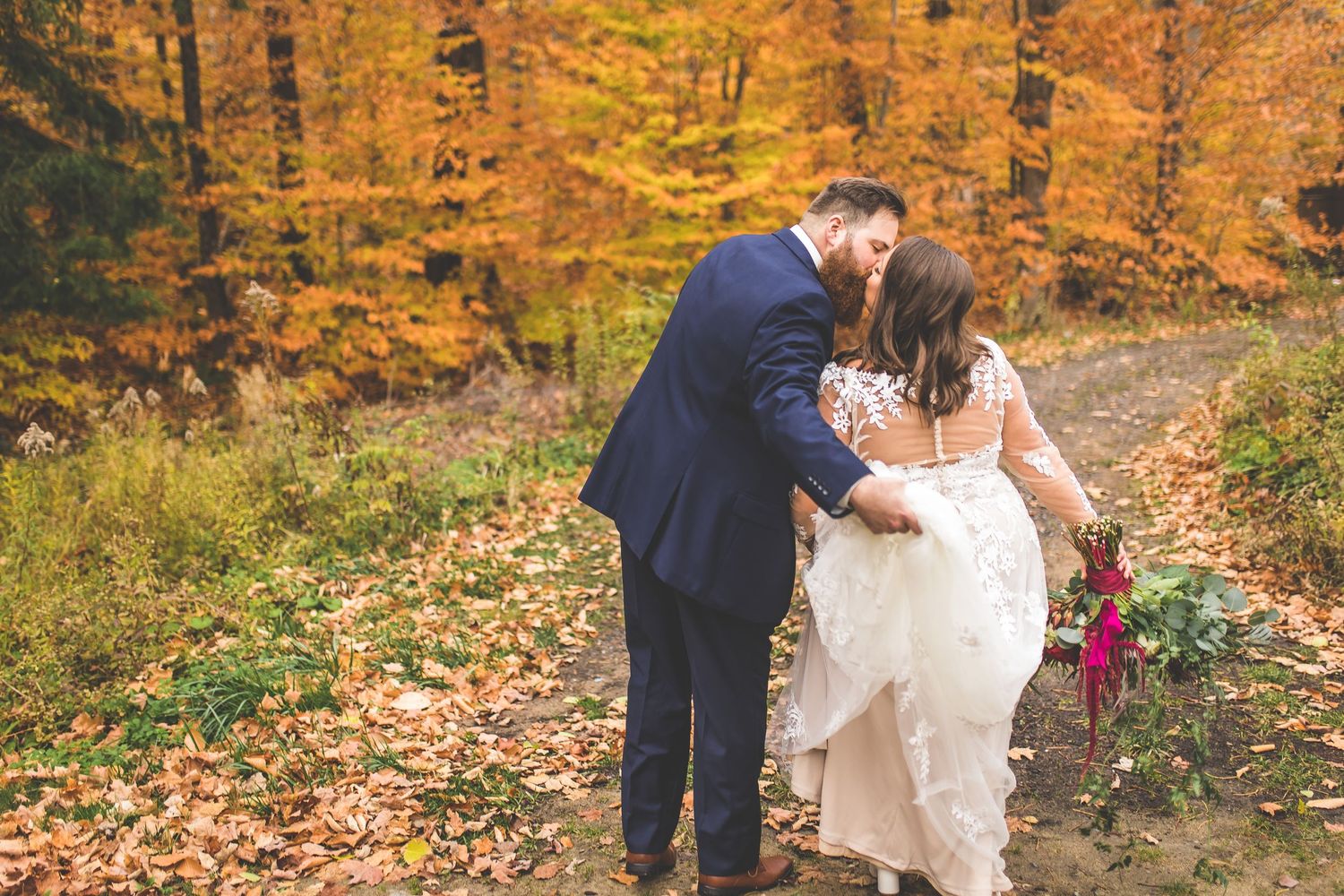 Wedding couple walks through fallen leaves in an autumn forest setting.