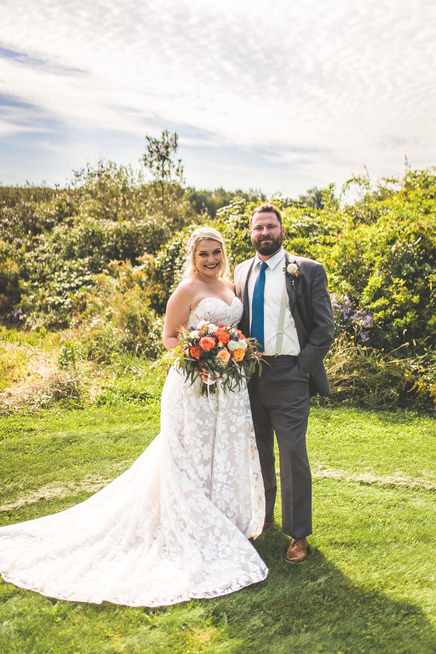 Bride in lace dress holds vibrant bouquet while posing with groom in outdoor garden setting.