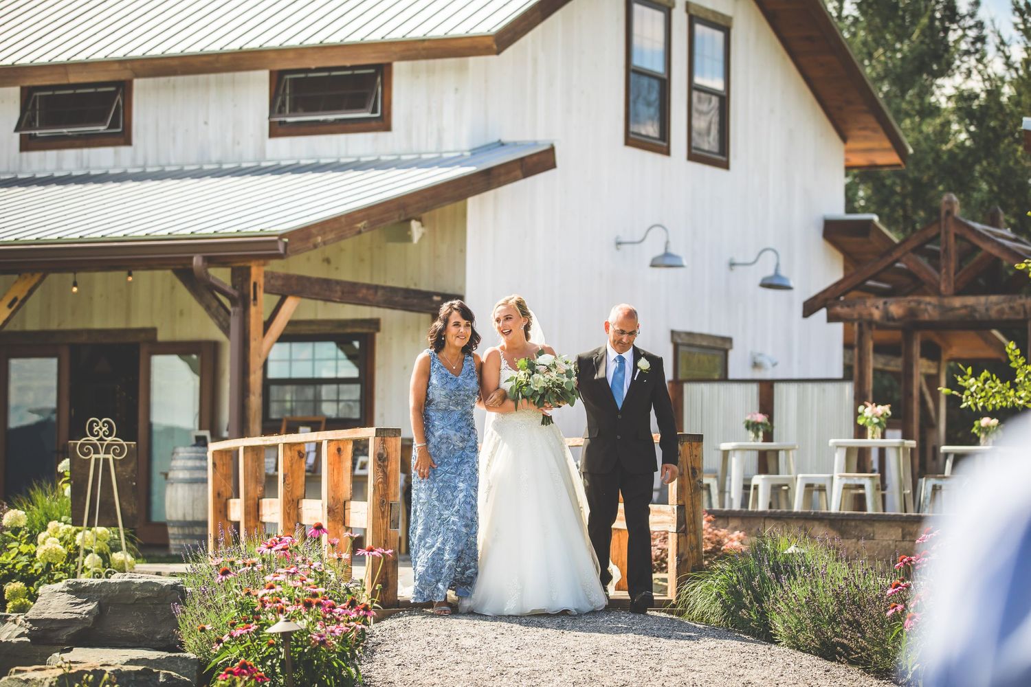 A wedding party walks down a garden path outside a rustic white farmhouse with blooming flowers.