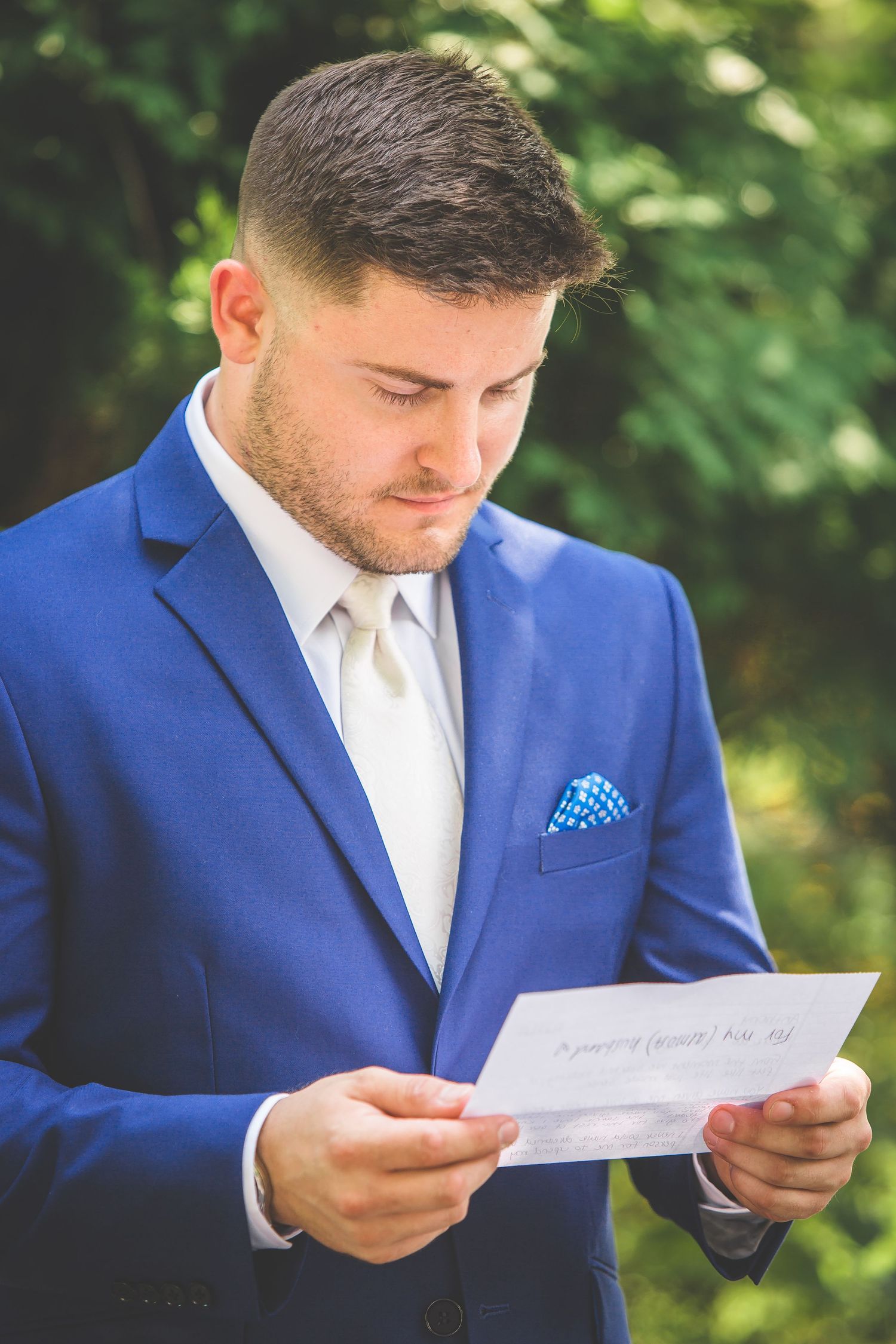 Groom in royal blue suit reading a letter outdoors before the ceremony.