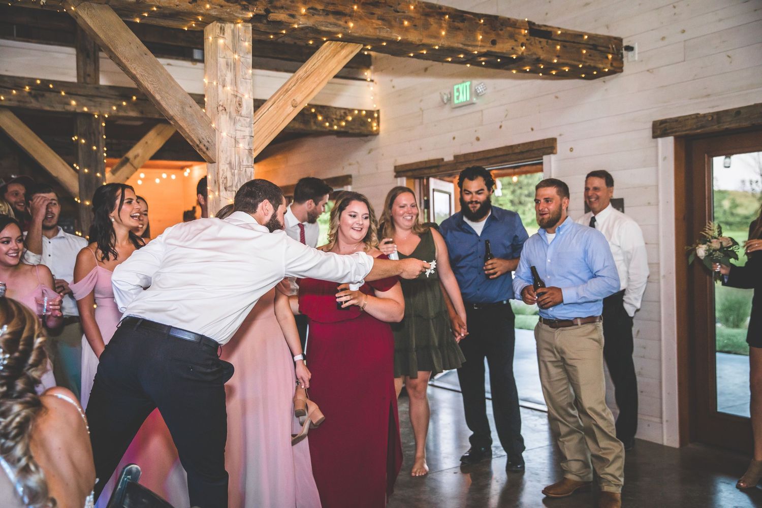 Wedding guests dancing in lines during reception under string lights and wooden beams.