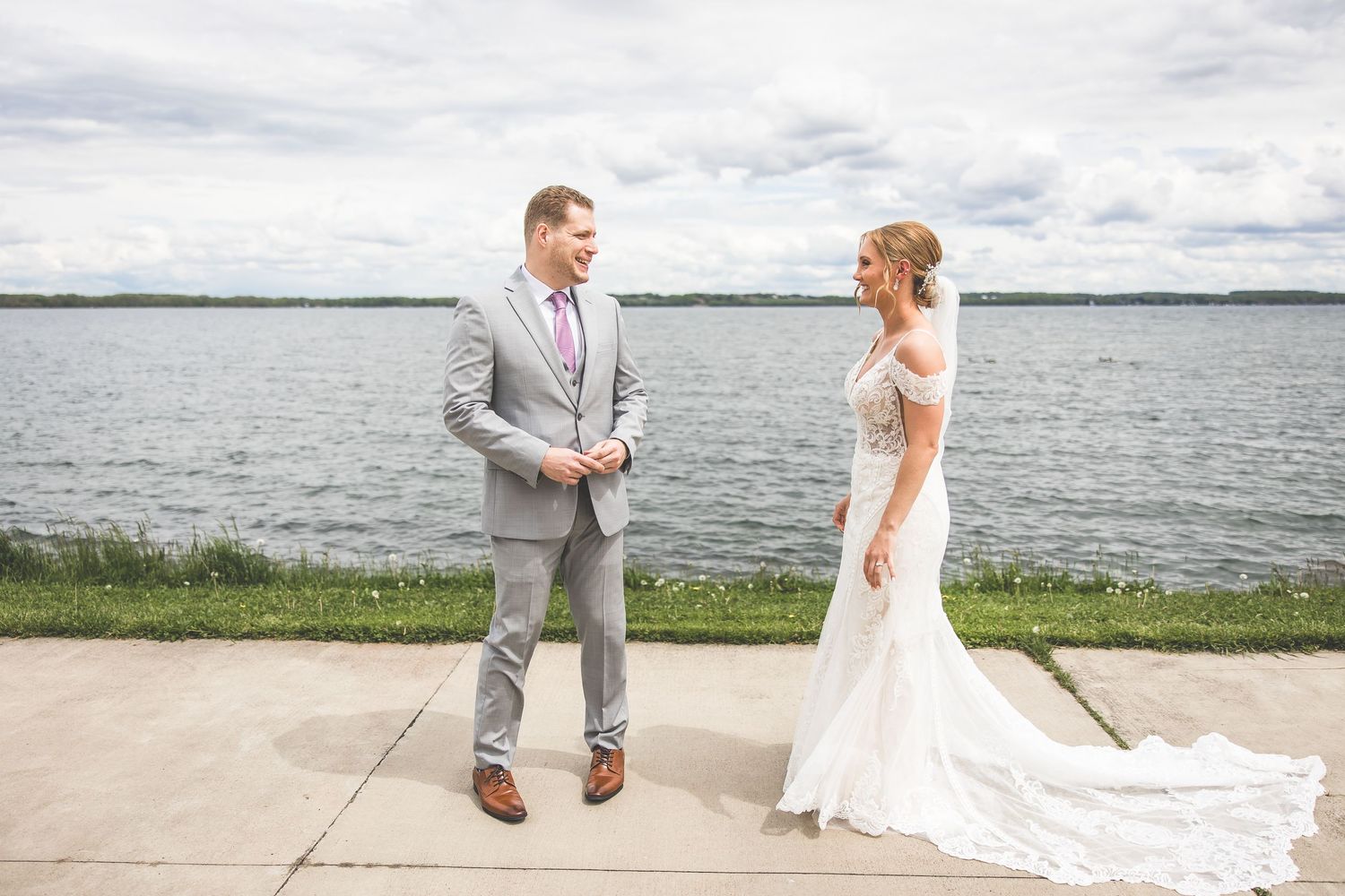 Couple sharing first look moment by waterfront with bride in lace gown and groom in grey suit.