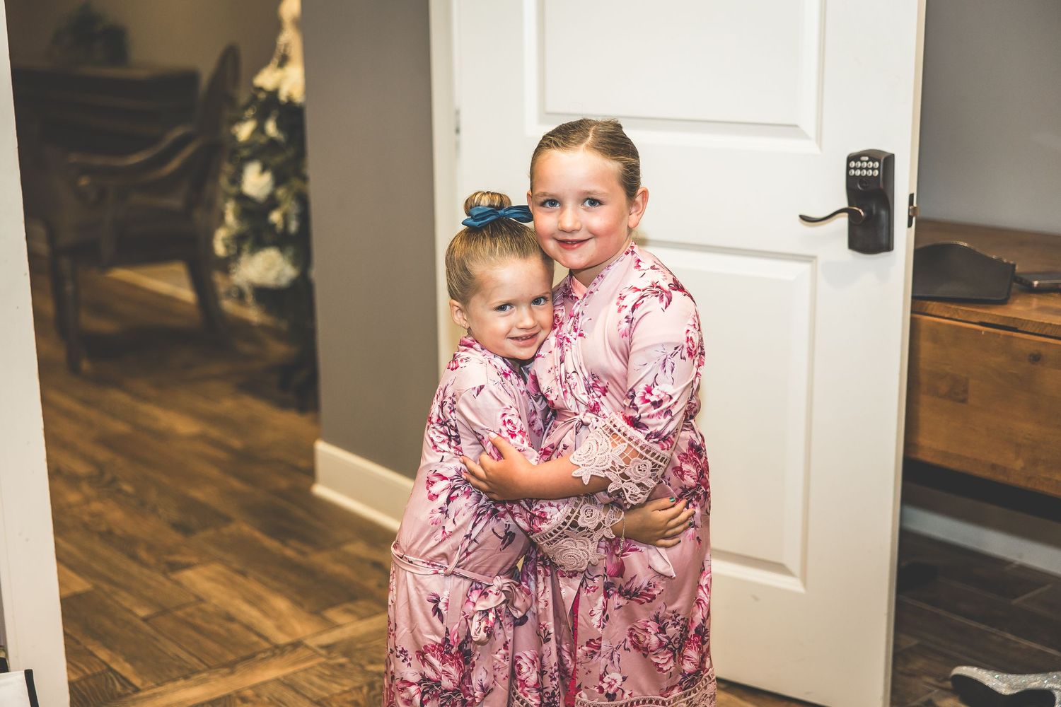 Two young attendants in matching pink floral dresses embrace during wedding preparations.