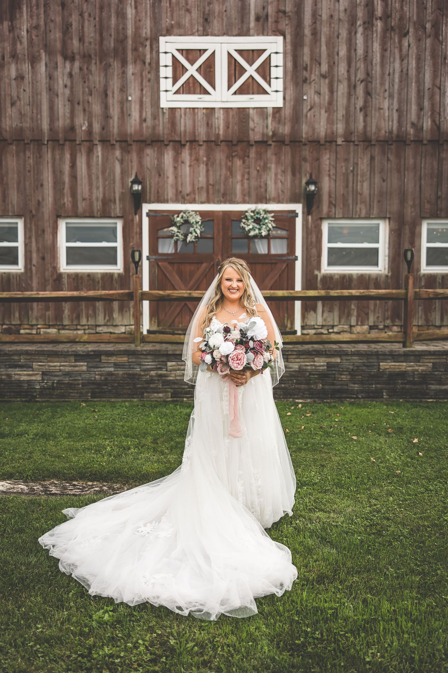 Bride in flowing white gown poses in front of rustic barn with floral bouquet.