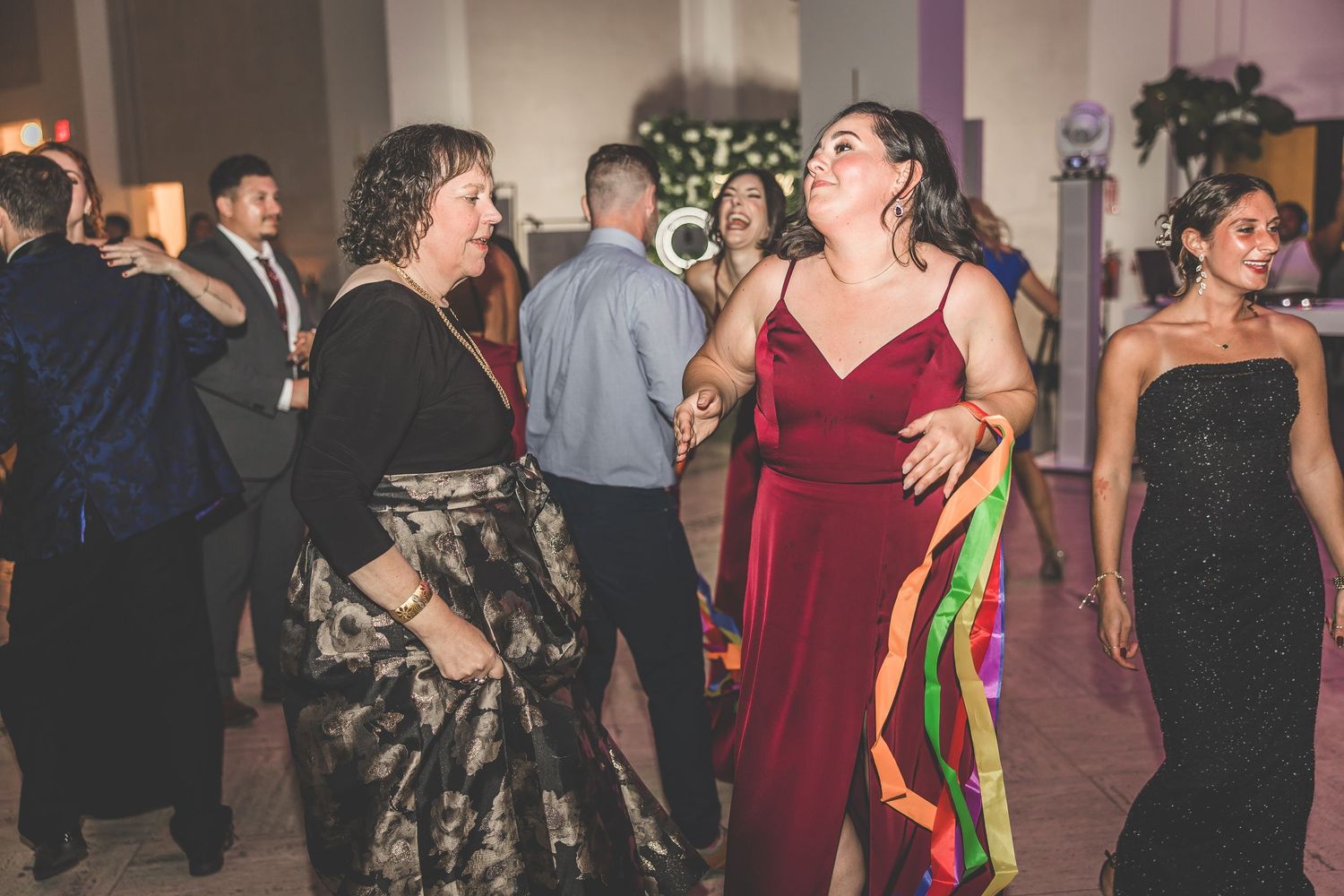 Wedding guests dance and celebrate on a dimly lit dance floor at an indoor reception venue.
