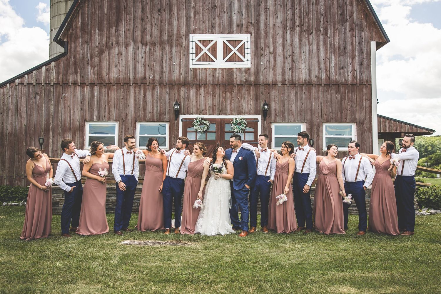Large wedding party lined up in front of a traditional barn wearing blue and brown attire.