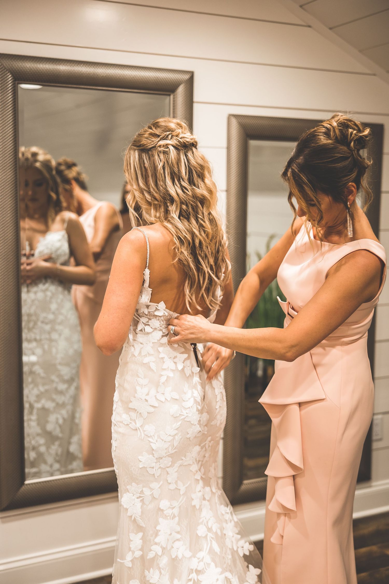 Helper assisting bride with lace wedding dress in front of mirrors.